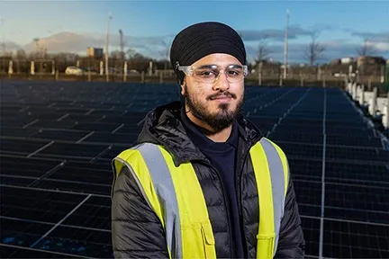 Young smiling man wearing black turban and work clothing, standing by solar panels