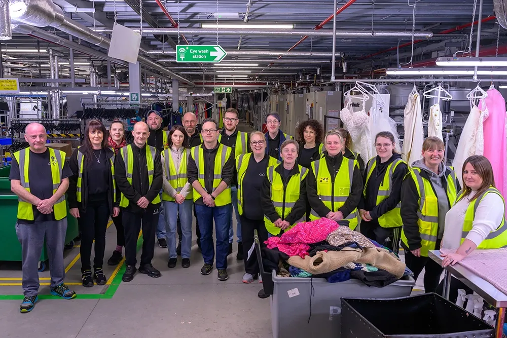 A group of people wearing hi-vis vests stand in a line. Various items of clothing are on coat hangers behind them.