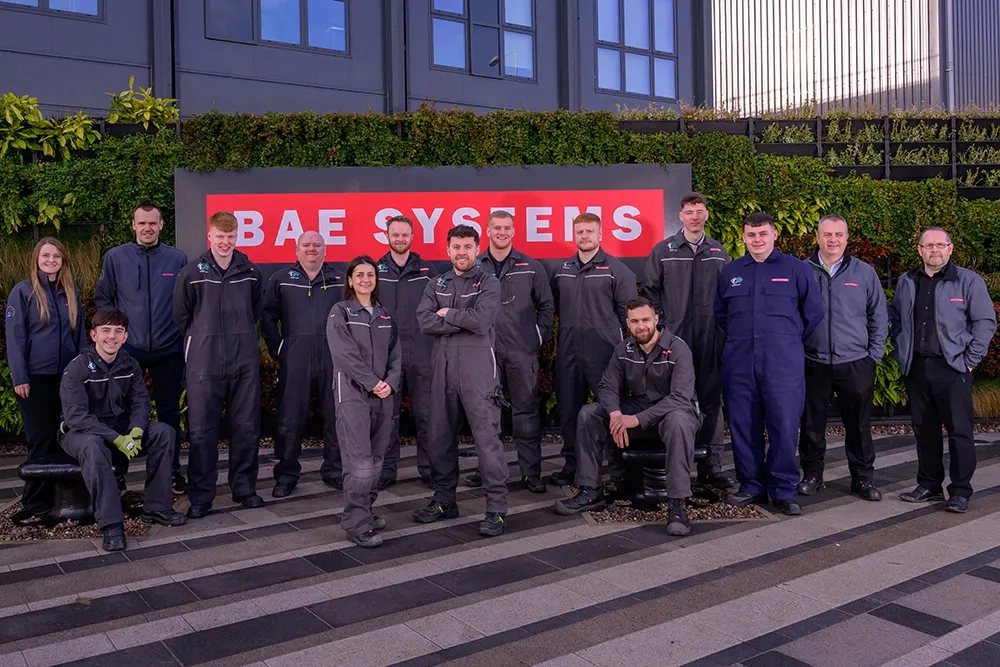 A group of people, all wearing boiler suits, stand in front of a red sign that says 'BAE Systems'.