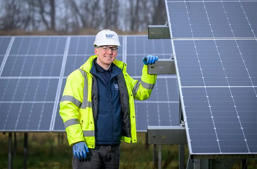 A young man standing next to a ground-mounted solar panel. He's wearing a hi-vis jacket, helmet and safety goggles.