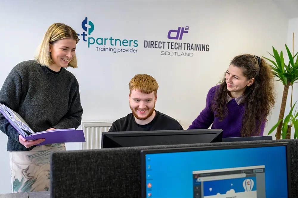 A young man sitting at a desk using a computer while two young women stand at either side of him. A wall sign in the background says 'Direct Tech Training Scotland'.