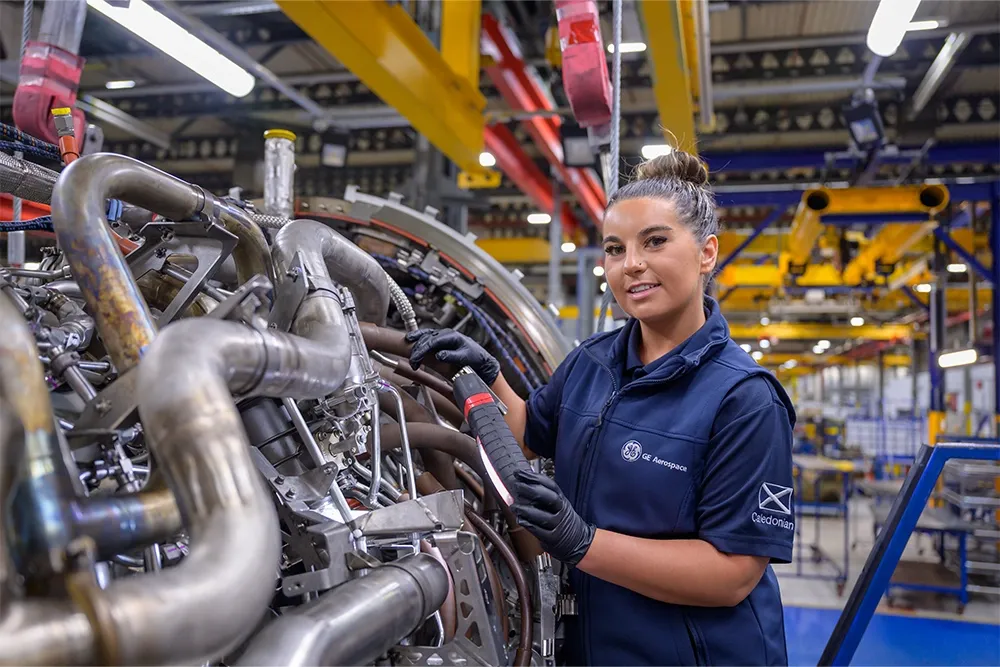 A young woman, wearing blue overalls, smiles at the camera while she carries out maintenance on an industrial machine.