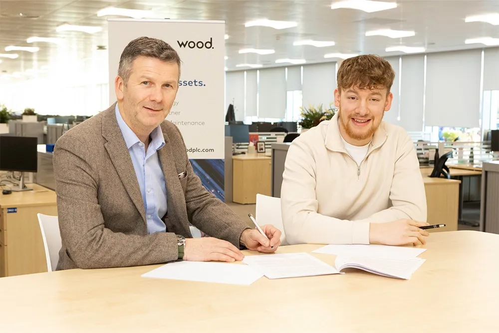 Two men sit at a desk and one of them uses a pen to fill out paperwork.